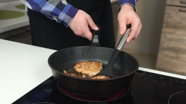 Gros homme dans un tablier frites côtelettes sur la cuisinière électrique en appuyant et en les retournant avec une spatule. Un mec épais dans un tablier fait cuire des escalopes sur le four électrique en les pressant et en les retournant avec une spatule .