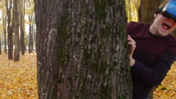 Facet w głowie zamontowany wyświetlacz krzyczy i chowa się za drzewem, człowiek w wirtualnej rzeczywistości zestaw słuchawkowy jest w jesiennym parku w słoneczny dzień — Wideo stockowe