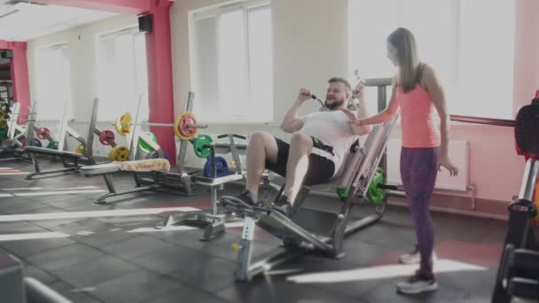 Guy Swing hans ben muskler på Training Simulator i gym med privat tränare. Viktminskning borrar från personlig individuell instruktör för fat man i Fitness Club. Tjock person som gör fysiska övningar — Stockvideo