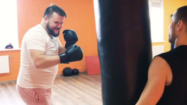 Indywidualny trening z osobistym trenerem. Pulchny facet w rękawiczkach bokserskich bije worek wykrawania w klubie fitness. Ćwiczenia odchudzania dla Grubego człowieka. Trening z instruktorem w siłowni. Prywatny trener grubych — Wideo stockowe