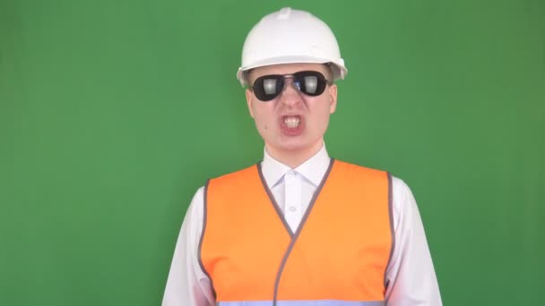 Angry caucasian boss in black glasses and a protective helmet and in a signal vest yelling at his subordinate and looking at the camera, green background, hromakey, industry — Stock Video