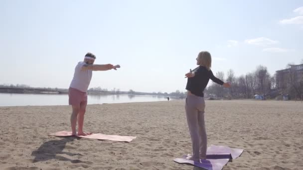 Instructor de yoga dinámico femenino caucásico enseña a un hombre completo con barba, clases de yoga en la naturaleza, playa de arena, río, mejora personal — Vídeos de Stock