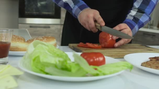 Buclatý chlápek vaří doma hamburgery v kuchyni. Muž krájí rajčata na hrací desce. Zdravý životní styl, smažené a škodlivé vysoce kalorické potraviny. Riziko obezity a nadváhy. Zpomaleně. — Stock video