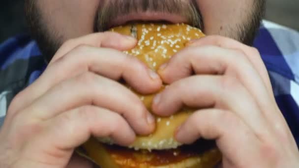 Closeup vousatý muž pokoušeje hamburger, který uvařil. Chlápek sní domácí jídlo. Zdravý životní styl, smažené a vysoce kalorické škodlivé potraviny. Riziko obezity a nadváhy. Zpomaleně. — Stock video