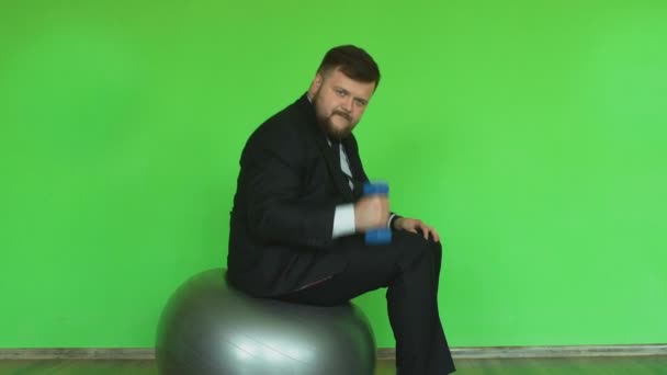 Funny Plump man i kostym med hantel sitter och rör sig på övning bollen i Fitness Club. Chromakey grön. Skäggiga tjocka killen i svart jacka, skjorta och slips är i gymmet — Stockvideo