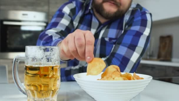 Крупный план лишний вес или толстяк ест чипсы с кружкой пива на кухне. Нездоровый образ жизни. Стеклянная кружка пильзенера или пива с болезненными картофельными чипсами. Алкогольный напиток и высокая калория — стоковое видео