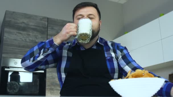 Fat man sitter i köket vid bordet och dricker skummande öl från mugg. En tjock kille i ett förkläde sitter vid bordet och klunkar skummande alkoholhaltiga dryck lager från en kopp. — Stockvideo