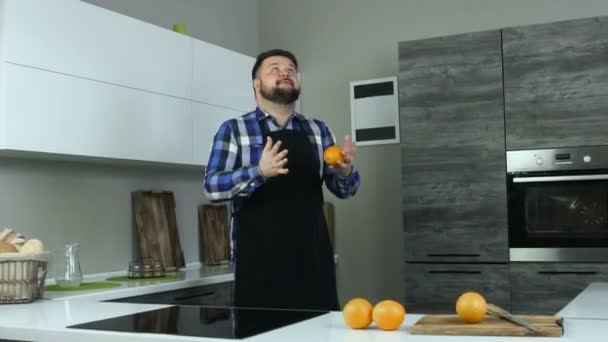 En fet man i ett förkläde i köket kastar upp och försöker jonglerar apelsiner, som sedan faller. En fyllig skäggiga kille med skägg jugglar frukter, som sedan släppa. — Stockvideo