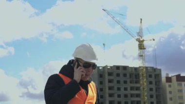 Kızgın mühendis ya da inşaatçı telefonda inşaat işlerinden endişeli bir şekilde bahsediyor. Beyaz kasklı, turuncu renkli yelekli ya da yelekli patron ya da şef astlarını azarlıyor ya da azarlıyor.