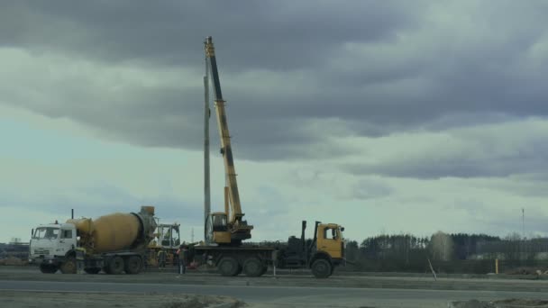 작업자는 도로를 따라 자동차 크레인으로 가로등이 있는 기둥을 설치합니다. 건설 기계, 콘크리트 또는 시멘트 믹서 수송 트럭은 작업 영역에 있습니다. 엔지니어는 고속도로에 가로등 기둥을 설치합니다. — 비디오