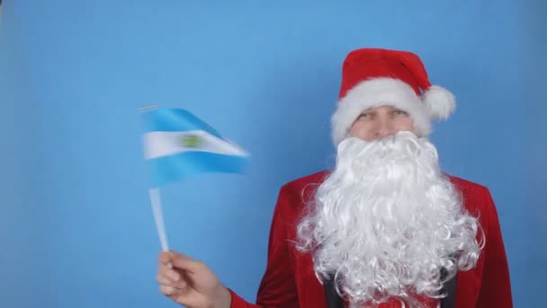 En man i jultomtedräkt med skägg som viftar med Argentinas flagga på blå bakgrund. Begreppet nyår semester i världen — Stockvideo
