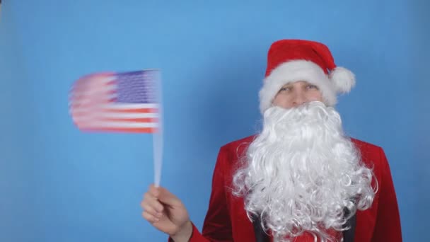 Un hombre disfrazado de Papá Noel con barba ondeando la bandera de Estados Unidos sobre un fondo azul. Concepto de vacaciones de Año Nuevo en el mundo — Vídeo de stock