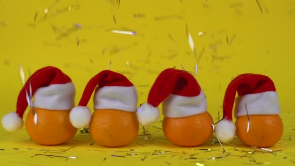 Confetti decorations falls on some mandarin oranges in a santa claus red hat. Concept of the new year or Christmas. Yellow background. Start of holidays and celebrations. — ストック動画