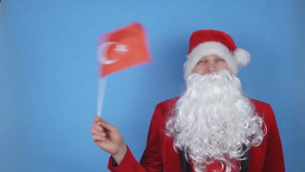 A man in a Santa Claus costume with a beard waving the flag of Turkey on a blue background. New Year holidays concept in the world — Stock Video
