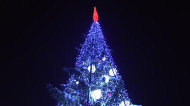 Çelenklerle süslenmiş Noel ağacının tepesinde geceleri ışıklar parlıyor. Kışın şehirde yeni yıl arifesi. Tatillerin ve kutlamaların başlangıcı kavramı. Geçmişi. Alt görünüm