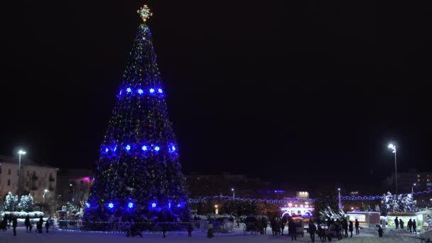 Árbol de Navidad decorado con guirnaldas y luces brilla por la noche en la plaza central de la ciudad. Nochevieja en la ciudad. La gente del pueblo camina y se relaja. Concepto de inicio de fiestas y celebraciones — Vídeo de stock