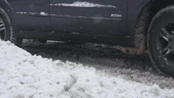 Автомобіль з зимовими шинами їде по сніговій дорозі в денний час у снігопаді. Колеса змиваються в повільному русі. Погані погодні умови для руху, хуртовина. Небезпека для поїздок. Скляний чорний лід на землі — стокове відео