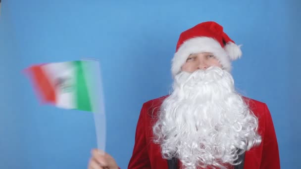 A man in a Santa Claus costume with a beard waving the flag of Mexico on a blue background. New Year holidays concept in the world — Stock Video