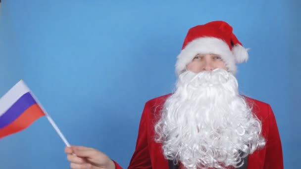 A man in a Santa Claus costume with a beard waving the flag of Russia on a blue background. New Year holidays concept in the world — Stock Video