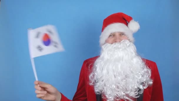 Een man in een kerstman kostuum met een baard zwaaiend met de vlag van Zuid-Korea op een blauwe achtergrond. Nieuwjaarsvakantie concept in de wereld — Stockvideo