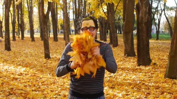 Giovane uomo caucasico in una maschera divertente gioisce in autunno e getta foglie gialle nel cielo, sfondo, rallentatore — Video Stock