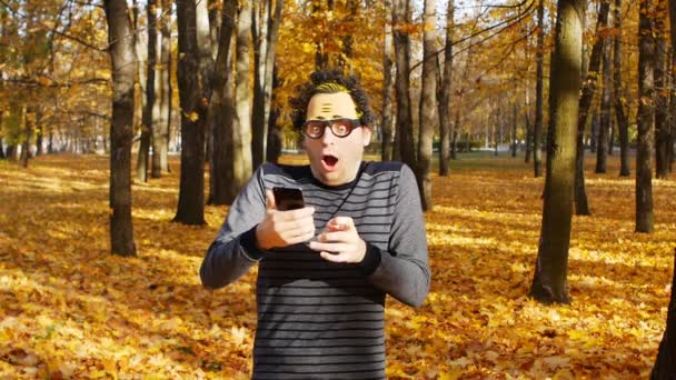 A young man in a wacky mask holds a smartphone in his hands against the backdrop of an autumn park and enjoys his win, luck, slow motion — Stock Video