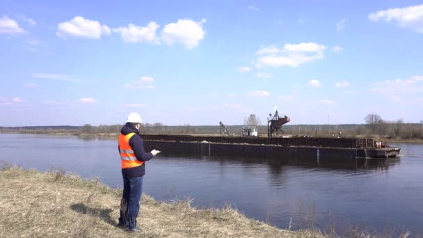 Ingénieur dans un gilet de signalisation sur le fond de la rivière sur laquelle se dresse un navire minier de sable de rivière — Video