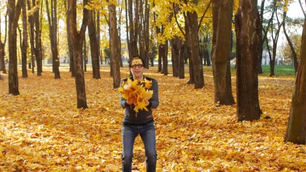 Mladý běloch v legrační masce se raduje na podzim a vyhazuje žluté listí do nebe, pozadí, zpomaluje — Stock video