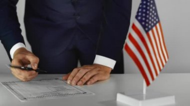 Birleşik Devletler başkanlık seçimlerinde oy veren bir adam. Adam kağıt oy pusulasını dolduruyor. Masada ABD bayrağı var. Takım elbiseli biri 2020, 2024 'te adaylar arasında seçim yapar. Genel oylama