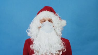 Noel, Yeni Yıl ve Coronavirus COVID konsepti. Koruyucu tıbbi maskeli Noel Baba gibi bir adam işaret parmağıyla yasak işareti gösterir. Mavi arkaplanda yasaklama hareketi