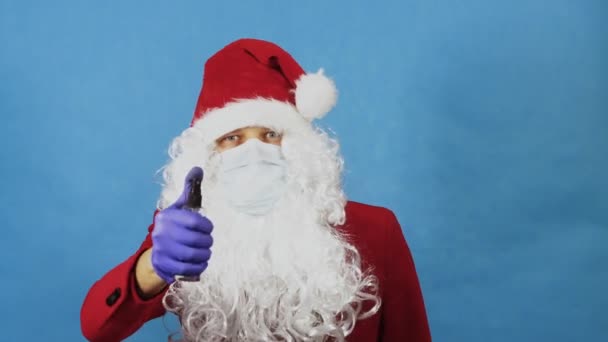 Mann wie ein Weihnachtsmann in medizinischer Schutzmaske, Handschuhe mit antiseptischem Desinfektionsspray. Coronavirus COVID und Weihnachten, Neujahr Konzept. Pandemie. Blauer Hintergrund — Stockvideo