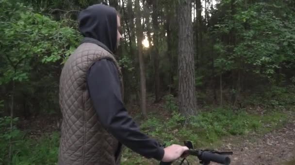 Человек в капюшоне прокатывает велосипед через лес на фоне солнечного заката, замедленного движения — стоковое видео