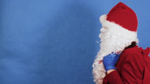 Noël, Nouvel An et Coronavirus COVID concept. Homme comme un Père Noël dans un masque médical de protection et des gants avec un sac de cadeaux sur son dos entre, regarde directement la caméra et part. Pandémie — Video