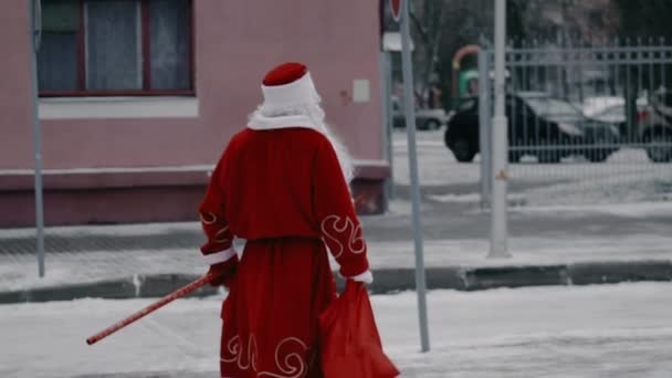 Rosyjski Ded Moroz jak Święty Mikołaj spaceruje po zimowym mieście, aby dać prezenty. Koncepcja Bożego Narodzenia i Nowego Roku. Szczupły facet z kijem — Wideo stockowe