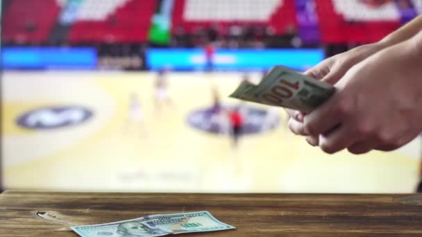 Um homem conta dólares no fundo de uma TV em que um torneio de basquete é mostrado. Conceito para casas de apostas e apostas esportivas, espaço de cópia para texto — Vídeo de Stock