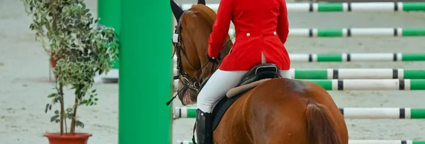 Horse horizontal banner for website header, poster, wallpaper. Rider in red and white uniform perfoming jump at show jumping competition. Riding hall and hurdle as a background.