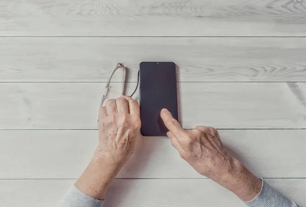 Old wrinkled hands holding mobile smart phone. Elderly woman using digital phone showing something on display, light table. Template, copy space mockup. Teaching, technologies for seniors concept.