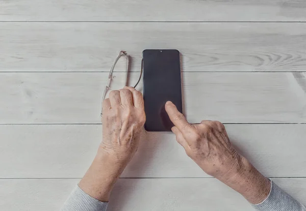 Old wrinkled hands holding mobile smart phone. Elderly woman using digital phone showing something on display, light table. Template, copy space mockup. Teaching, technologies for seniors concept.
