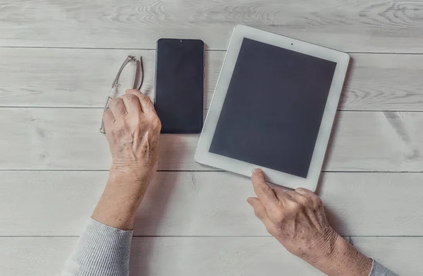 Old wrinkled hands, mobile smart phone and tablet. Elderly woman using a phone and tablet showing something on display, light table. Copy space mockup. Teaching, technology, seniors concept.