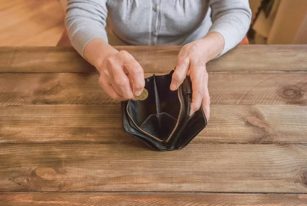 Old wrinkled hand holding coin, empty wallet, wooden background. Elderly woman throws a coin into a purse, counting. Saving money for future plan and retirement fund, pension, poorness, need concept.