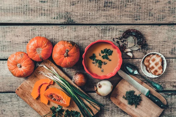 Pumpkin soup on wooden table, linen cloth, vintage cutlery. Autumn cream soup, seeds and parsley. Sliced herbs, pumpkin on cutting board, cooking process. Thanksgiving dinner, flat lay, top view.