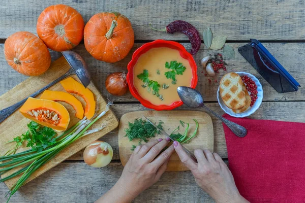 Pumpkin soup on wooden table, vintage. Woman hands, herbs, cooking process. Girl looking for a meal recipe on mobile phone while cooks at home. Thanksgiving, vegetarian, autumn. Flat lay, top view.