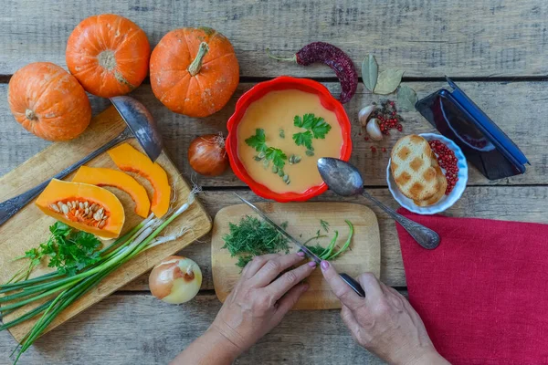 Pumpkin soup on wooden table, vintage. Woman hands, herbs, cooking process. Girl looking for a meal recipe on mobile phone while cooks at home. Thanksgiving, vegetarian, autumn. Flat lay, top view.