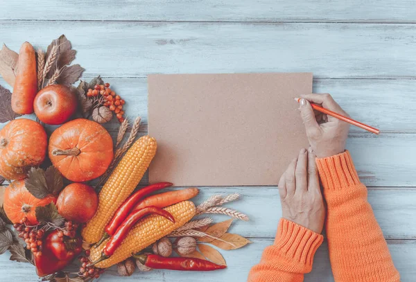 Autumn background, fallen leaves, fruits, vegetables on rustic wooden table. Woman hands with seasonal corner set, aged paper, copy space. Thanksgiving healthy and fresh food, top view, flat lay.