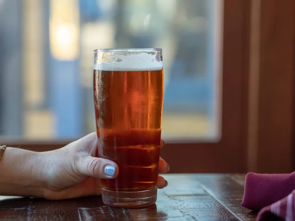 Kvinna s hand med rosa naglar gripande en bärnstensfärgad öl glas framför fönstret — Stockfoto
