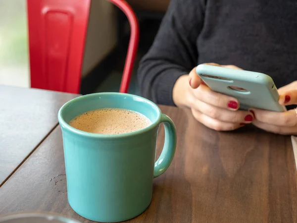 Mujer usando el teléfono mientras espera a que el chocolate caliente se enfríe en la taza de té — Foto de Stock