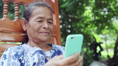 happy asian senior mature woman relaxing and using smartphone, speaking on mobile phone with family, thai asia elderly grandmother enjoy smart outdoor lifestyle with wireless technology device