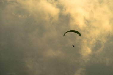 Gün batımında bulutlu gökyüzünde yükselen paraglider silueti.