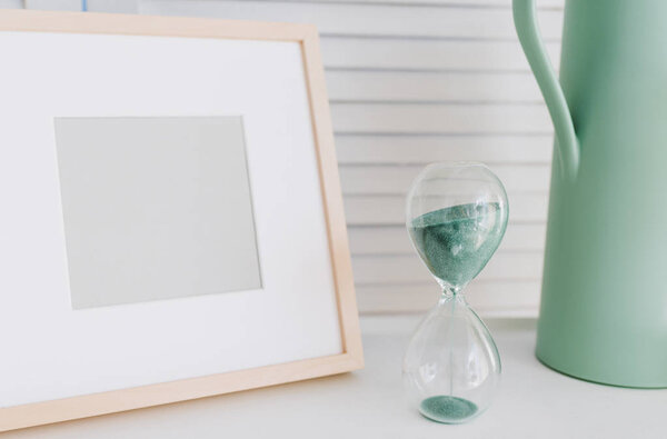 Пустой фотокаркас, поливающий горшок и часы со стеклом или песком в качестве элементов интерьера перед белой стеной. Концепция времени
.