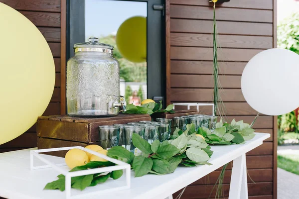 Φρέσκα ολόκληρα λεμόνια με φύλλα σε ένα λευκό τραπέζι κοντά σε ένα βάζο με λεμονάδα και μπαλόνια σε εξωτερικούς χώρους — Φωτογραφία Αρχείου
